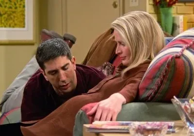 Homem moreno deitado em sofá verde conversa com mulher loira sentada, que olha para longe. Cenário de sala aconchegante.