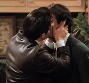 Dois homens se beijam, um de jaqueta de couro e outro de suéter. Cenário natalino com guirlanda. Uma mulher os observa à esquerda.