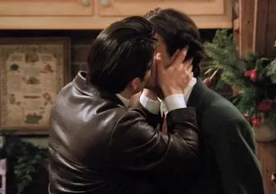 Dois homens se beijam, um de jaqueta de couro e outro de suéter. Cenário natalino com guirlanda. Uma mulher os observa à esquerda.