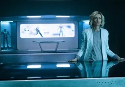 Mulher loira em jaqueta clara num laboratório futurista. Ao fundo, uma armadura e diagramas tecnológicos em telas iluminadas de azul.