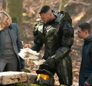 Um soldado blindado, uma mulher e um homem jovem examinam papéis e um caixote em uma floresta. Clima sério e futurista.