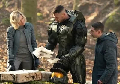 Um soldado blindado, uma mulher e um homem jovem examinam papéis e um caixote em uma floresta. Clima sério e futurista.