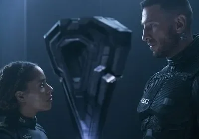 Uma mulher e um homem em uniformes futuristas olham um objeto geométrico misterioso. Cenário sci-fi escuro com luz azul.