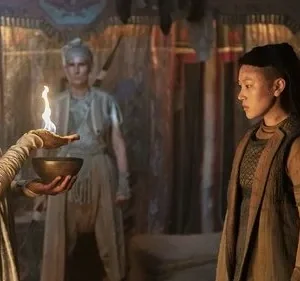 Três mulheres em trajes místicos. Uma segura uma tigela com fogo, observada por outra. Cenário escuro e ritualístico.