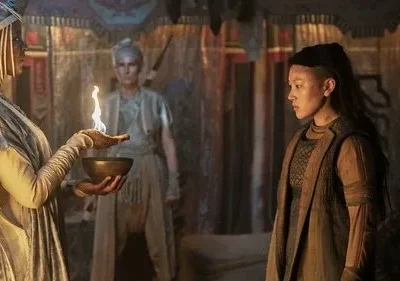 Três mulheres em trajes místicos. Uma segura uma tigela com fogo, observada por outra. Cenário escuro e ritualístico.