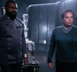 Um homem barbudo e uma mulher, ambos em trajes futuristas escuros com insígnias, num cenário tecnológico sombrio.