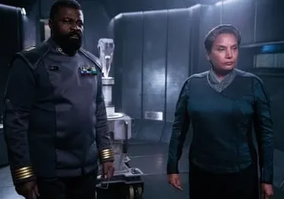 Um homem barbudo e uma mulher, ambos em trajes futuristas escuros com insígnias, num cenário tecnológico sombrio.