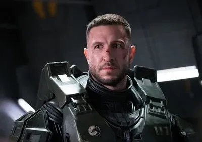 Homem sério com rosto machucado e armadura verde-escura futurista em ambiente metálico e escuro. Clima de ação sci-fi.