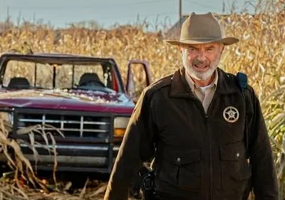 Xerife idoso de chapéu e barba em campo de milho seco. Ao fundo, um caminhão vermelho danificado. Clima rural e tenso.