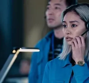 Jovem mulher asiática de cabelo claro e camisa azul, com headset, fala séria em uma sala de controle escura.