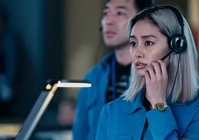Jovem mulher asiática de cabelo claro e camisa azul, com headset, fala séria em uma sala de controle escura.