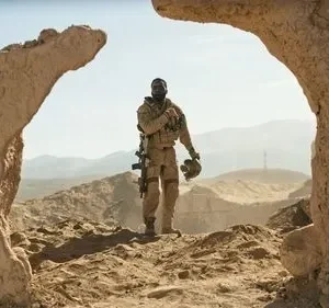 Um soldado negro em uniforme deserto, com rifle e capacete, em pé sob um céu azul, entre ruínas de argila e montanhas no deserto.