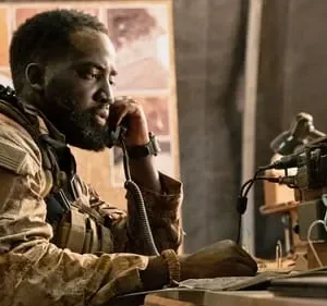 Um soldado negro barbudo em uniforme camuflado fala ao telefone, em um ambiente tático com mapas e equipamentos.