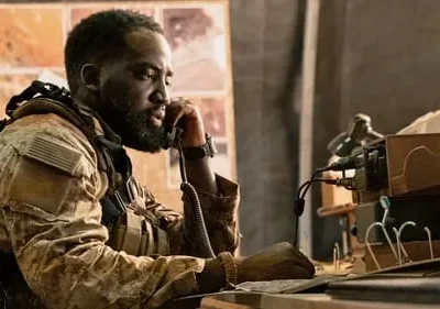 Um soldado negro barbudo em uniforme camuflado fala ao telefone, em um ambiente tático com mapas e equipamentos.