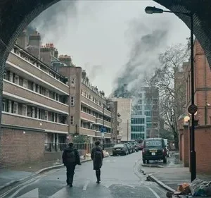Duas figuras de costas caminham por uma rua urbana sombria, sob um arco de tijolos. Fumaça densa sobe ao longe entre prédios, num cenário desolador.