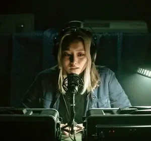 Mulher loira séria com fones de ouvido fala ao microfone em estúdio escuro. Uma luz pontual ilumina o rosto, criando um clima tenso.