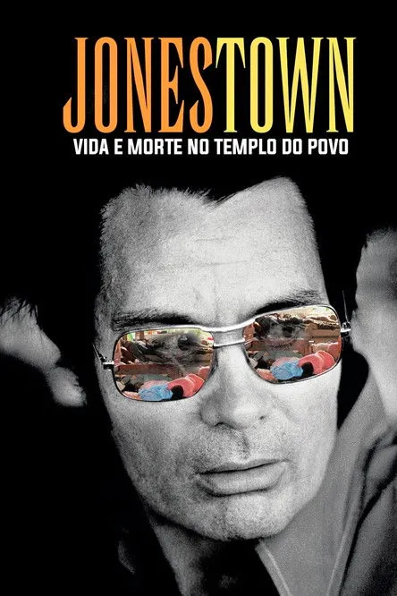 Jonestown:Vida e Morte no Templo do Povo 1 Close-up P&B de homem sério com óculos aviador. As lentes espelham figuras coloridas em vermelho e azul,em cena confusa e perturbadora.