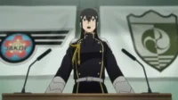Kaiju No. 8 – T01E05:Alistamento!8 Pessoa de uniforme militar escuro com detalhes dourados,boca aberta,em pódio com dois microfones. Bandeiras com emblemas ao fundo.