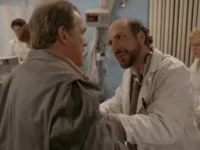 Um médico barbudo com estetoscópio conversa com um homem de casaco cinza em um hospital, com equipe médica ao fundo. Clima de diálogo sério.