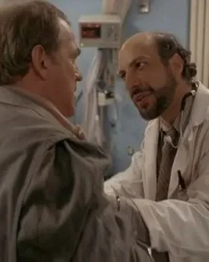 Um médico barbudo com estetoscópio conversa com um homem de casaco cinza em um hospital, com equipe médica ao fundo. Clima de diálogo sério.