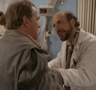 Um médico barbudo com estetoscópio conversa com um homem de casaco cinza em um hospital, com equipe médica ao fundo. Clima de diálogo sério.