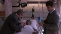 Homem acamado em hospital. Um homem mais velho o conforta, segurando a mão, enquanto outro homem de sobretudo faz anotações.