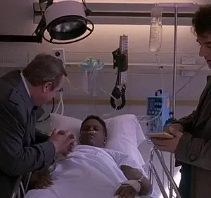 Homem acamado em hospital. Um homem mais velho o conforta, segurando a mão, enquanto outro homem de sobretudo faz anotações.