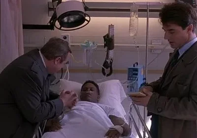 Homem acamado em hospital. Um homem mais velho o conforta, segurando a mão, enquanto outro homem de sobretudo faz anotações.