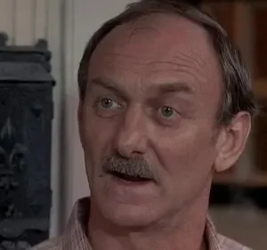 Homem maduro de bigode e cabelo grisalho, com olhar atento, em ambiente interno. Fundo mostra caixa ornamental escura.