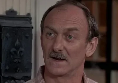 Homem maduro de bigode e cabelo grisalho, com olhar atento, em ambiente interno. Fundo mostra caixa ornamental escura.