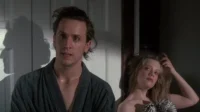 Um homem de roupão com cabelo bagunçado e ar pensativo ao lado de uma mulher loira, com olhar relaxado e sensual. Cena íntima.