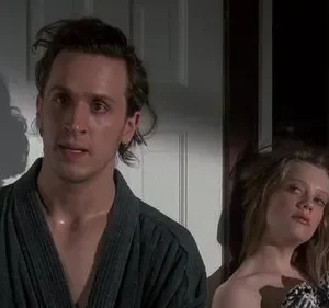 Um homem de roupão com cabelo bagunçado e ar pensativo ao lado de uma mulher loira, com olhar relaxado e sensual. Cena íntima.