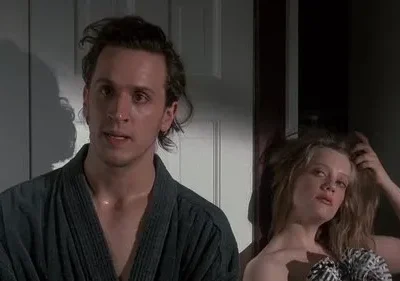 Um homem de roupão com cabelo bagunçado e ar pensativo ao lado de uma mulher loira, com olhar relaxado e sensual. Cena íntima.