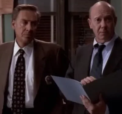 Dois homens de terno com expressões sérias em um escritório. O da direita segura uma pasta aberta. Clima formal.