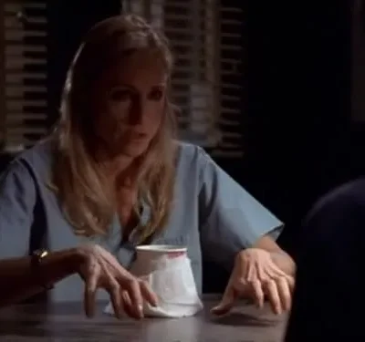 Mulher loira de scrubs azuis, com expressão tensa e mãos crispadas na mesa escura. Clima dramático.