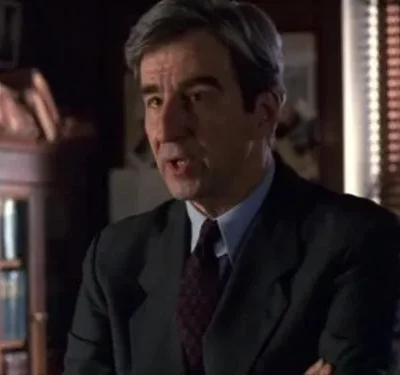 Homem de terno escuro e cabelo grisalho, com expressão séria, falando em um escritório com estantes.