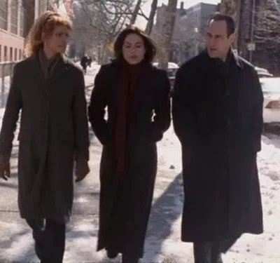 Três adultos (2 mulheres, 1 homem) de casacos de inverno caminham sérios em uma rua nevada da cidade.