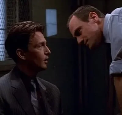 Dois homens em cena tensa. Um de terno olha para o outro de camisa, que se inclina sobre ele em atitude confrontadora.