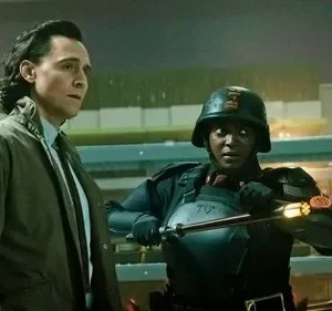 Homem em casaco marrom e mulher com uniforme tático e capacete, segurando bastão luminoso. Cenário de armazém, clima tenso.