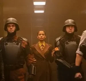Grupo de cinco pessoas em um corredor mal iluminado. Inclui uma mulher de uniforme futurista e um homem com gola eletrônica, ladeados por guardas.