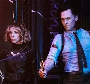 Uma mulher de armadura e um homem de terno, com adagas e expressões sérias. Fundo escuro com iluminação roxa e laranja dramática.