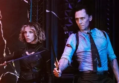 Uma mulher de armadura e um homem de terno, com adagas e expressões sérias. Fundo escuro com iluminação roxa e laranja dramática.