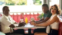 Três pessoas em um diner retrô. Um homem mais velho fala animadamente, enquanto um jovem e uma mulher de óculos observam com expressões sérias.