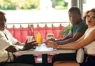 Três pessoas em um diner retrô. Um homem mais velho fala animadamente, enquanto um jovem e uma mulher de óculos observam com expressões sérias.