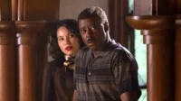 Um homem negro e uma mulher de cabelos escuros parecem assustados em um ambiente com painéis de madeira. O clima é tenso.