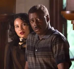 Um homem negro e uma mulher de cabelos escuros parecem assustados em um ambiente com painéis de madeira. O clima é tenso.