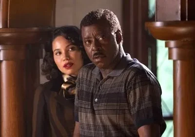 Um homem negro e uma mulher de cabelos escuros parecem assustados em um ambiente com painéis de madeira. O clima é tenso.