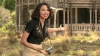 Jovem sorridente com câmera e roupa vintage gesticula, convidando para uma casa antiga ornamentada em meio à natureza.