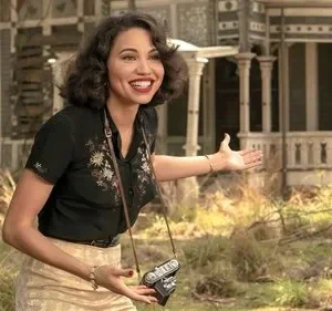 Jovem sorridente com câmera e roupa vintage gesticula, convidando para uma casa antiga ornamentada em meio à natureza.