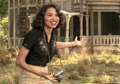 Jovem sorridente com câmera e roupa vintage gesticula, convidando para uma casa antiga ornamentada em meio à natureza.